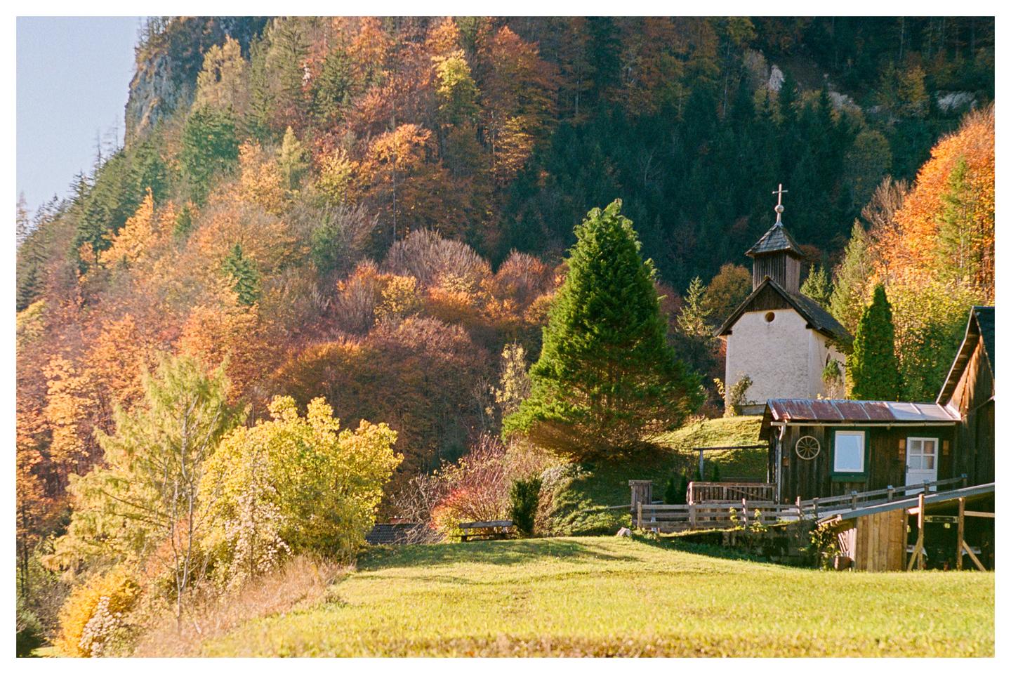 A small chapel and a wooden cabin are nestled on a grassy hill surrounded by a forest with autumn foliage.