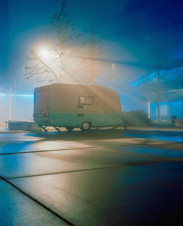 A caravan is parked on a paved surface at night, illuminated by a bright light source and surrounded by bare trees.
