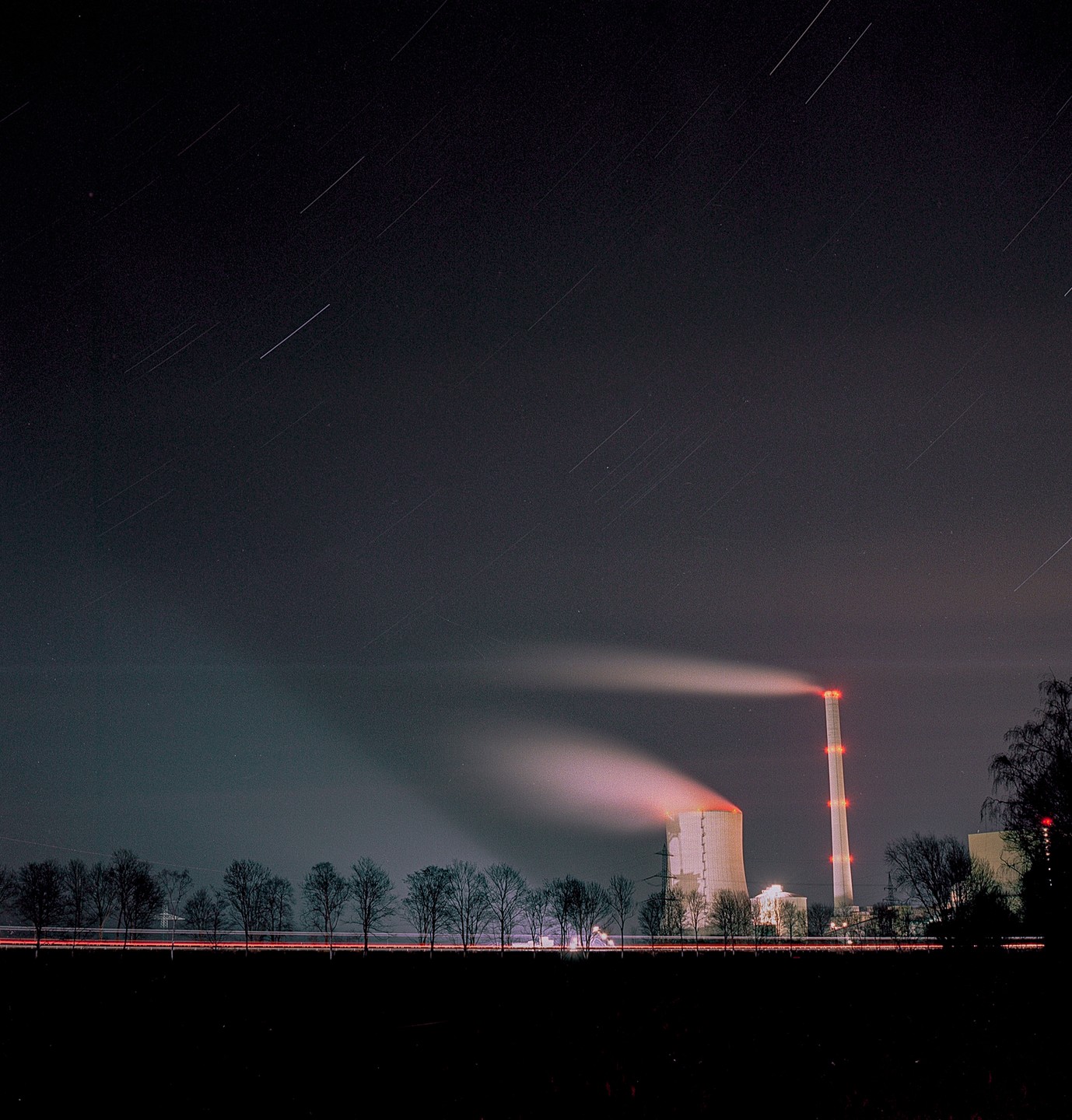 A power plant with two smoking chimneys stands under a night sky with star trails.
