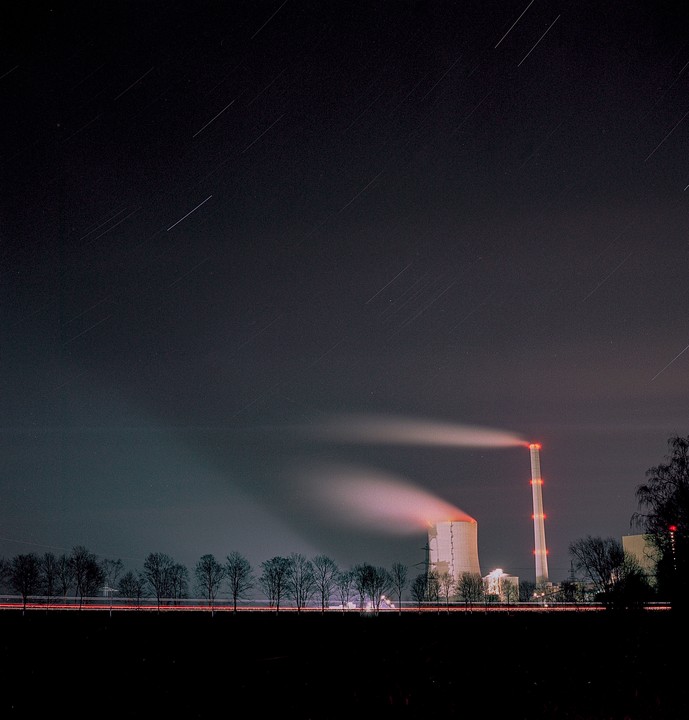 A power plant with two smoking chimneys stands under a night sky with star trails.