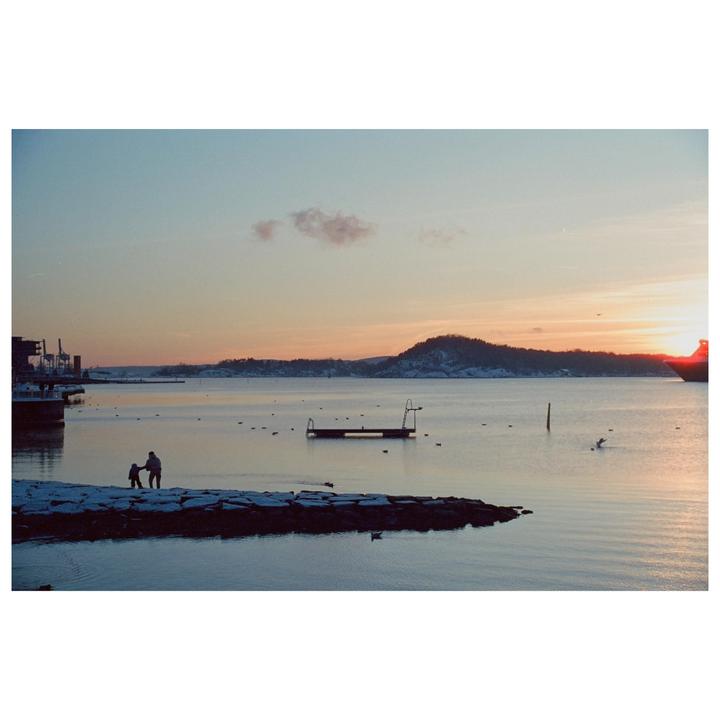 A man and child stand on a snowy pier overlooking a body of water with a sunset in the background.