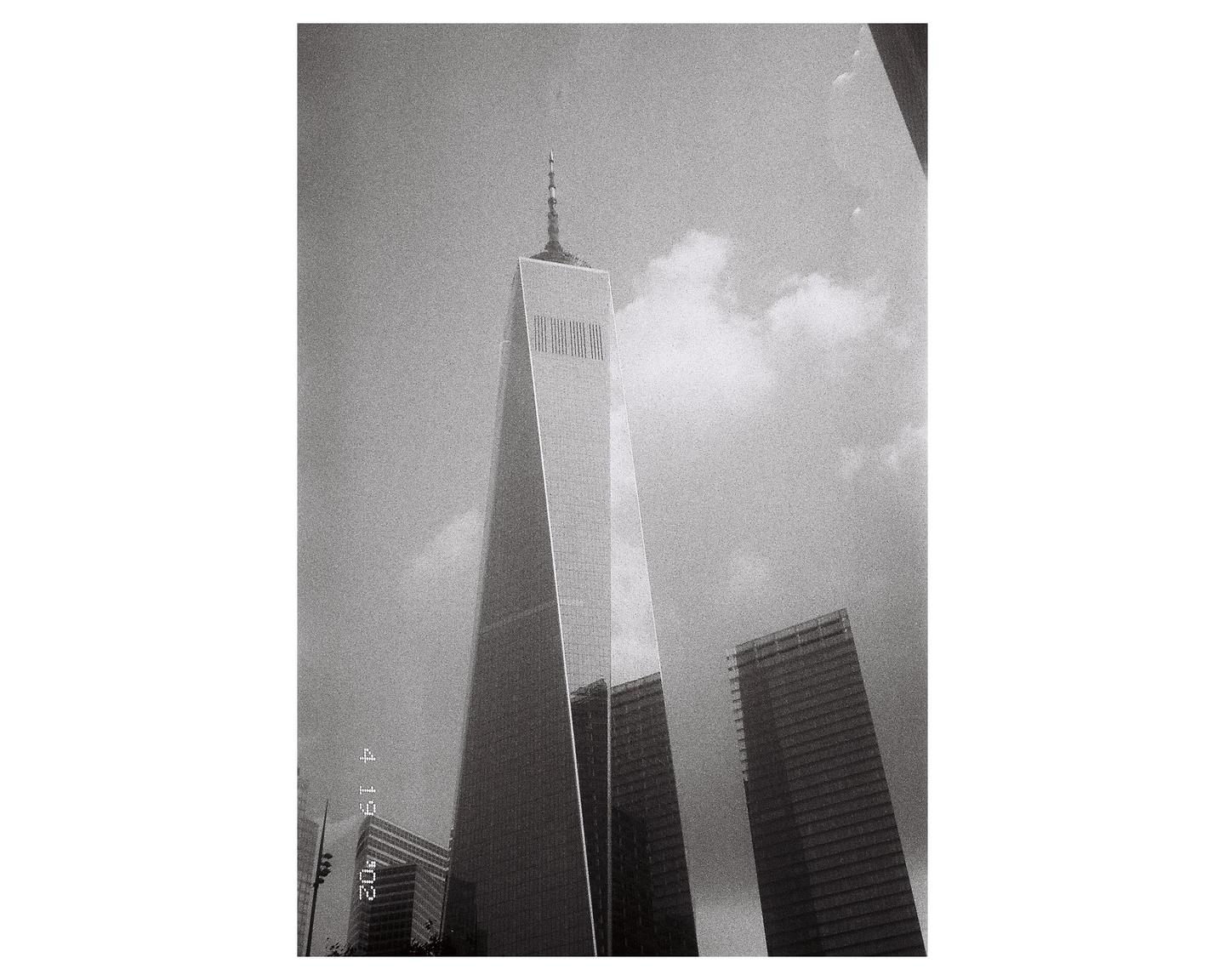 A low-angle view shows a tall, modern skyscraper with a spire against a cloudy sky, with other buildings visible in the background.