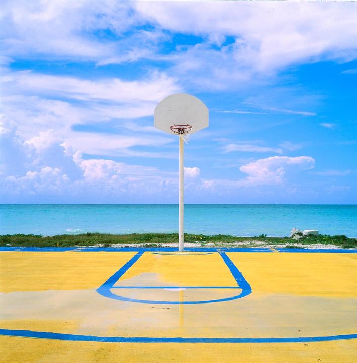 A basketball hoop stands on a yellow and blue court next to a turquoise ocean under a blue sky with clouds.