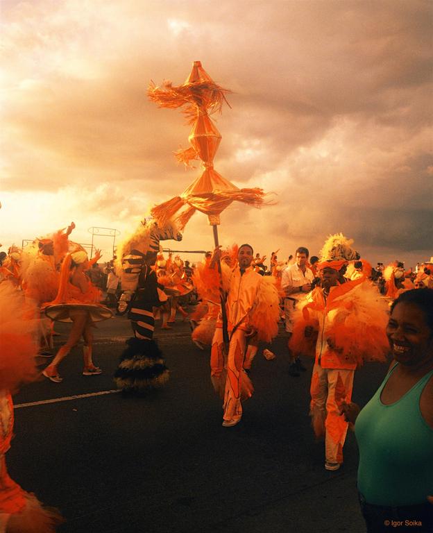A crowd of people in orange costumes are parading down a street under a dramatic sky.