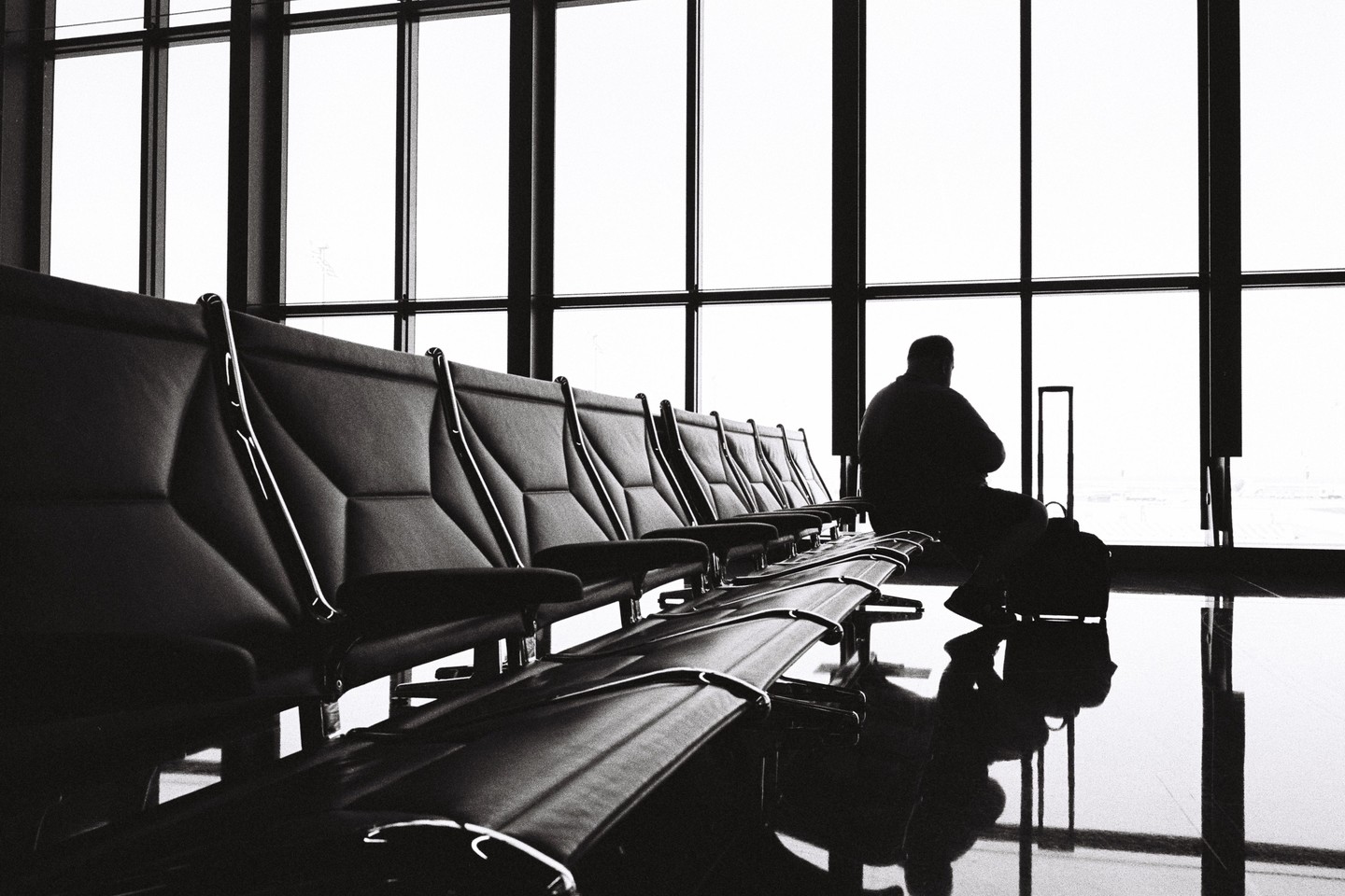 A man sits in silhouette in an airport waiting area with a row of empty seats and large windows.