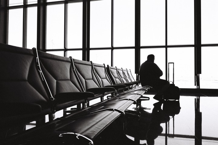 A man sits in silhouette in an airport waiting area with a row of empty seats and large windows.