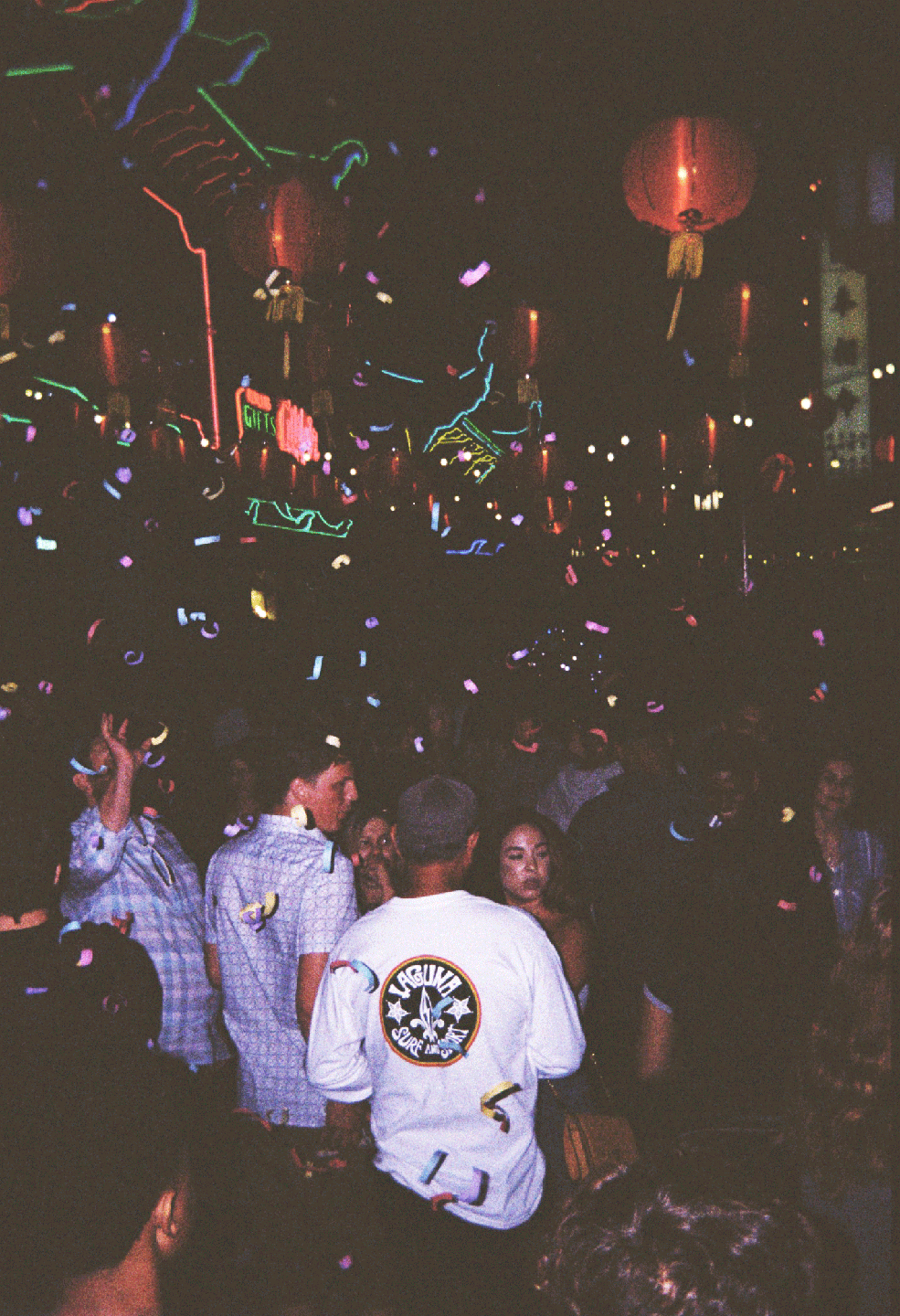 Confetti falls over a crowd of people at night with neon lights and red lanterns in the background.