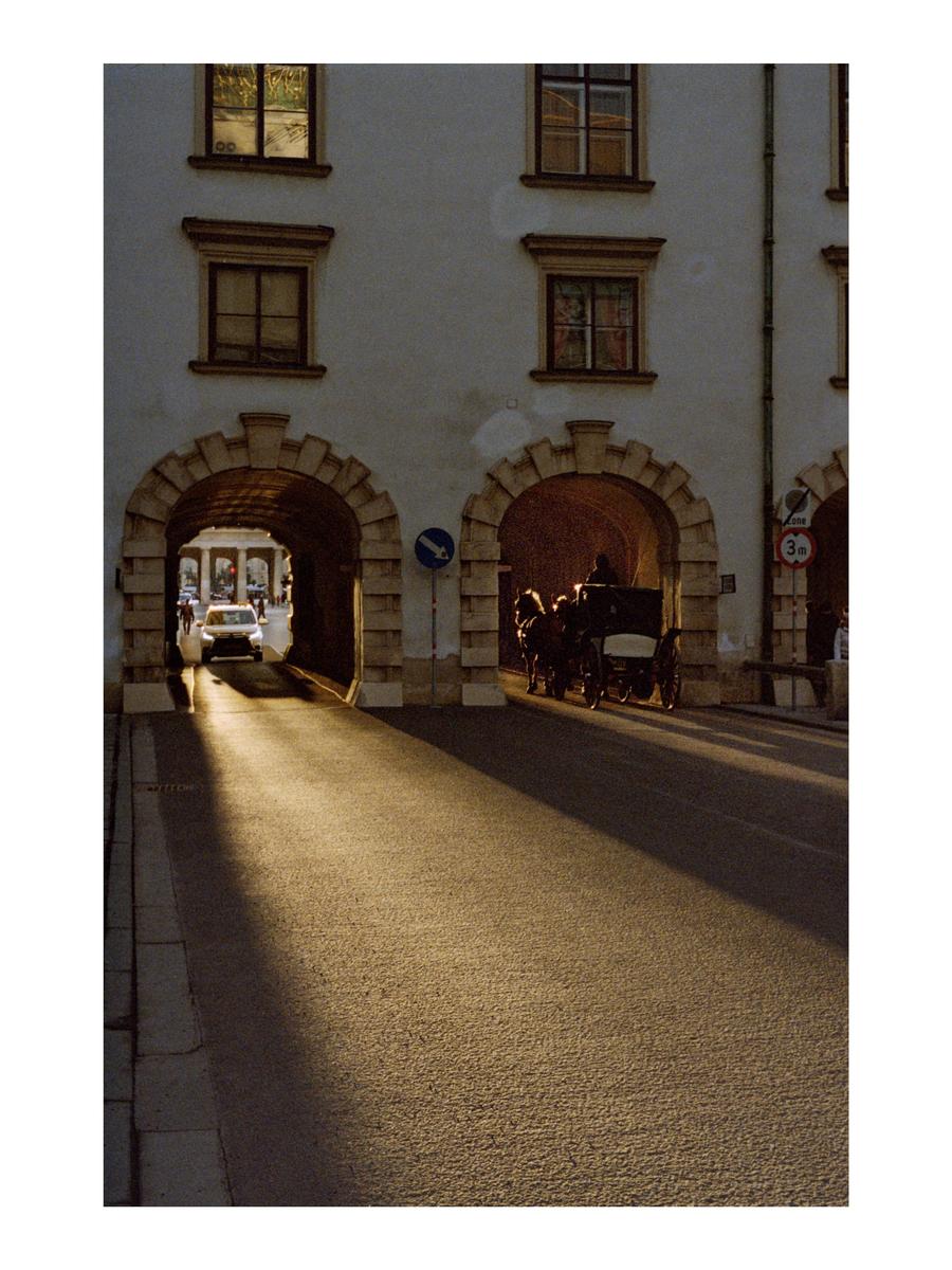 A horse-drawn carriage and a car are visible through two archways in a building facade, with a long shadow cast across the street.