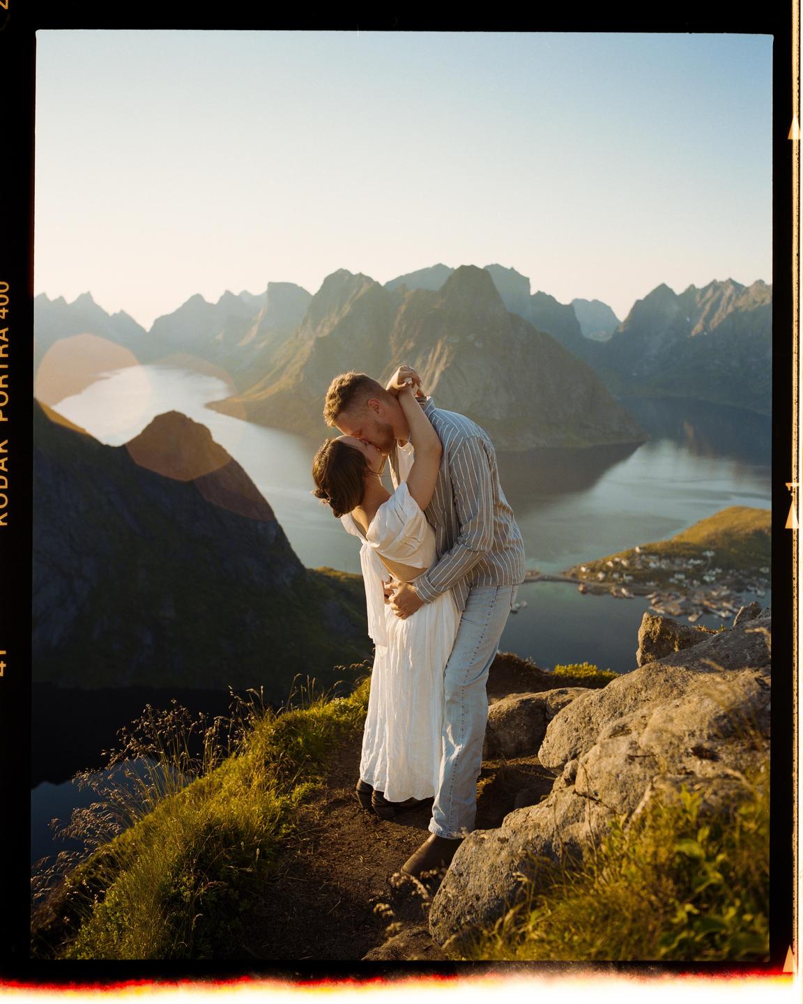 A couple kisses on a mountain overlooking a fjord and a small village.