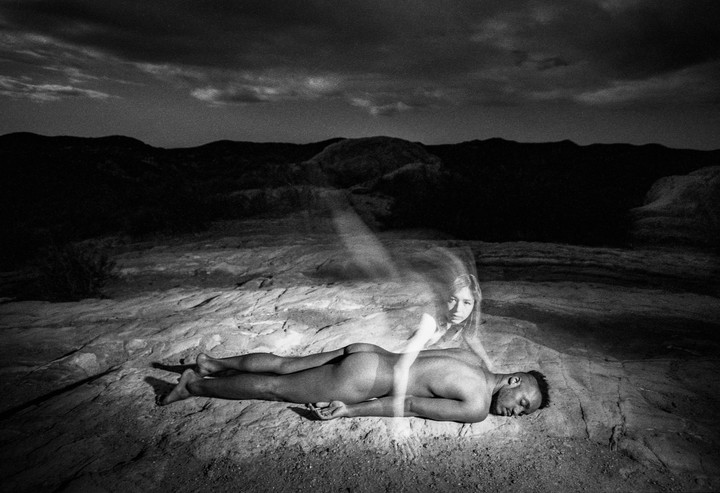 A man lies on the ground with a ghostly woman hovering above him in a desert landscape at night.