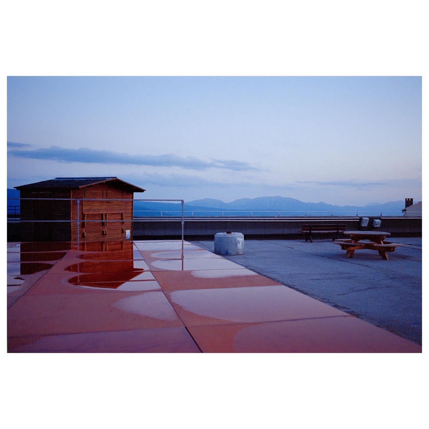 A wooden shed and a picnic table sit on a rooftop with mountains in the distance under a blue sky.