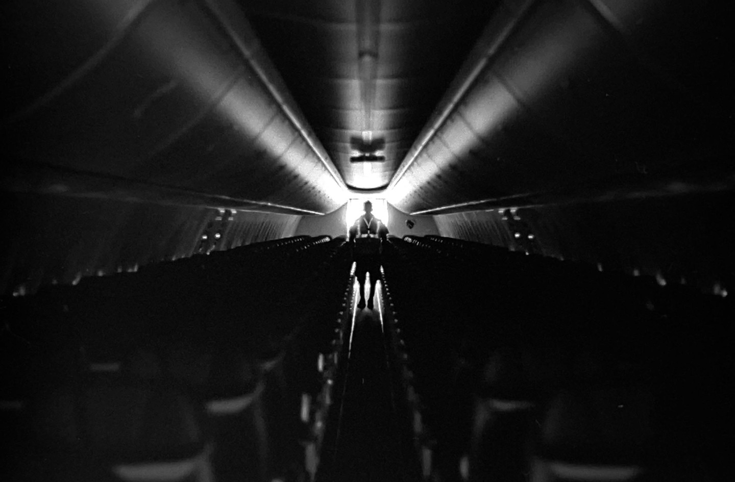 A person stands at the end of an airplane aisle, silhouetted by light.
