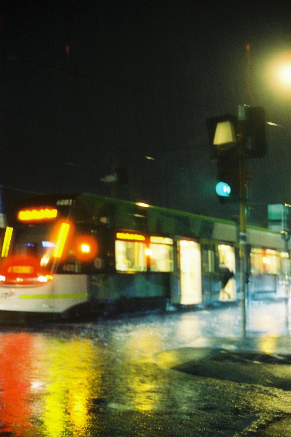 A tram with illuminated windows is stopped at a street corner at night, with reflections of its lights on the wet ground.
