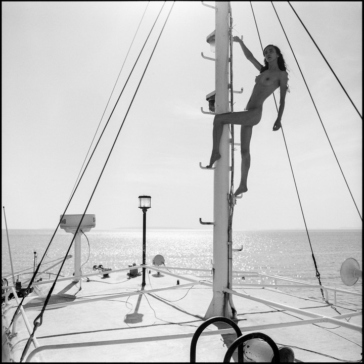 A nude woman climbs a ladder on a boat with the ocean in the background.