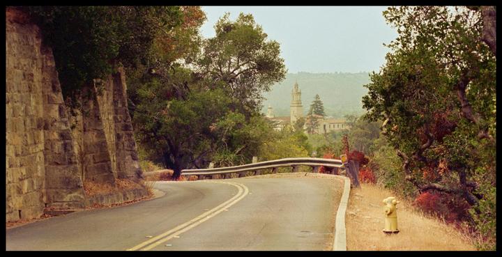 A winding road with a stone wall on the left leads to a distant building with a prominent tower.
