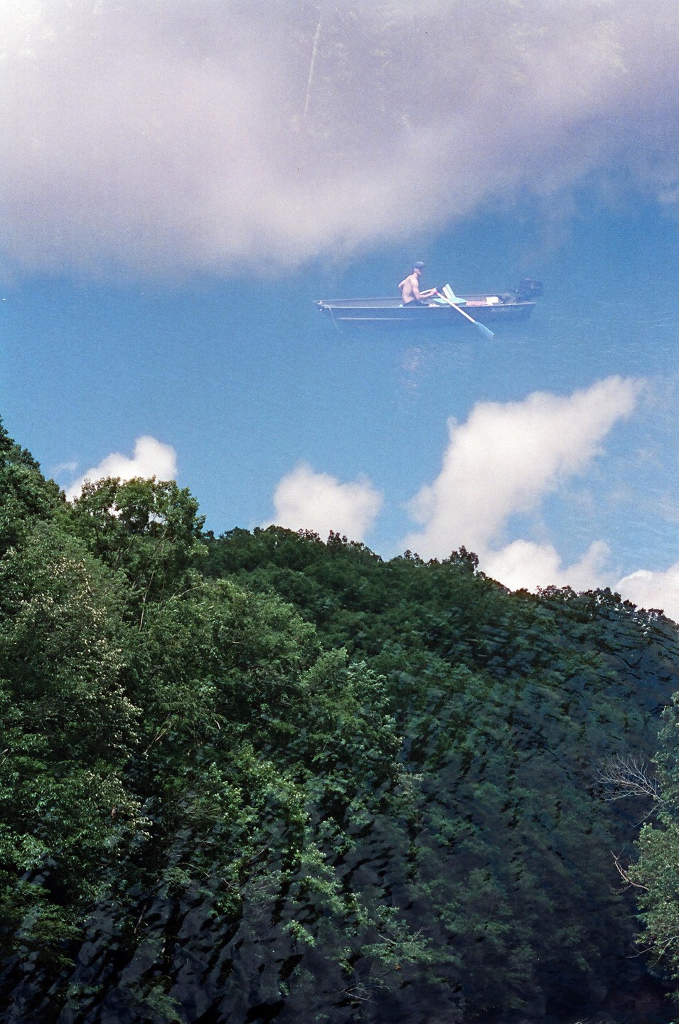 A man rows a small boat on a body of water with a forested hill in the foreground.