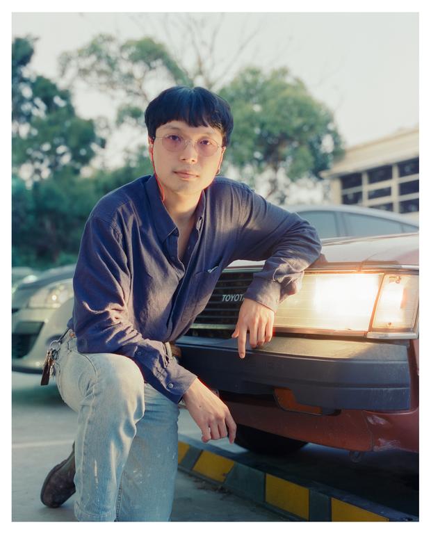 A man wearing glasses and a blue shirt kneels next to a car with his arm resting on the hood.