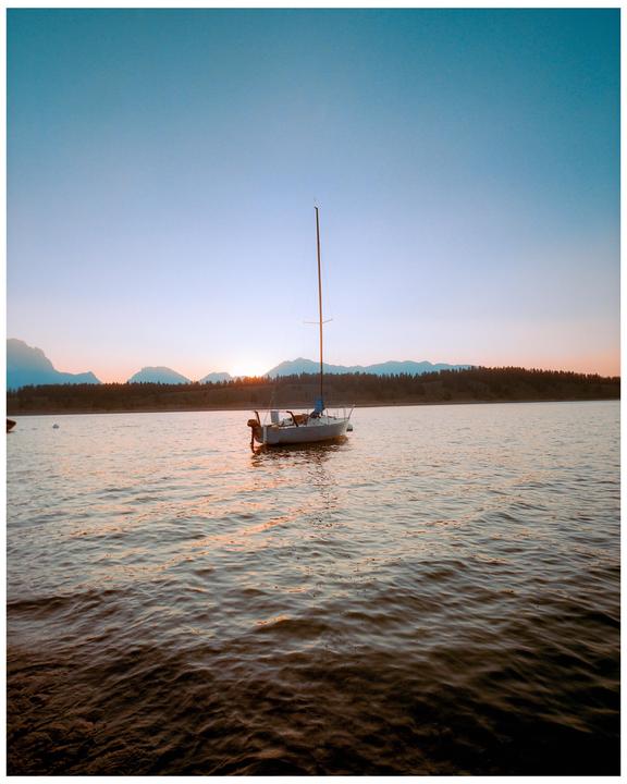 A sailboat floats on a lake with mountains and a forest in the background at sunset.