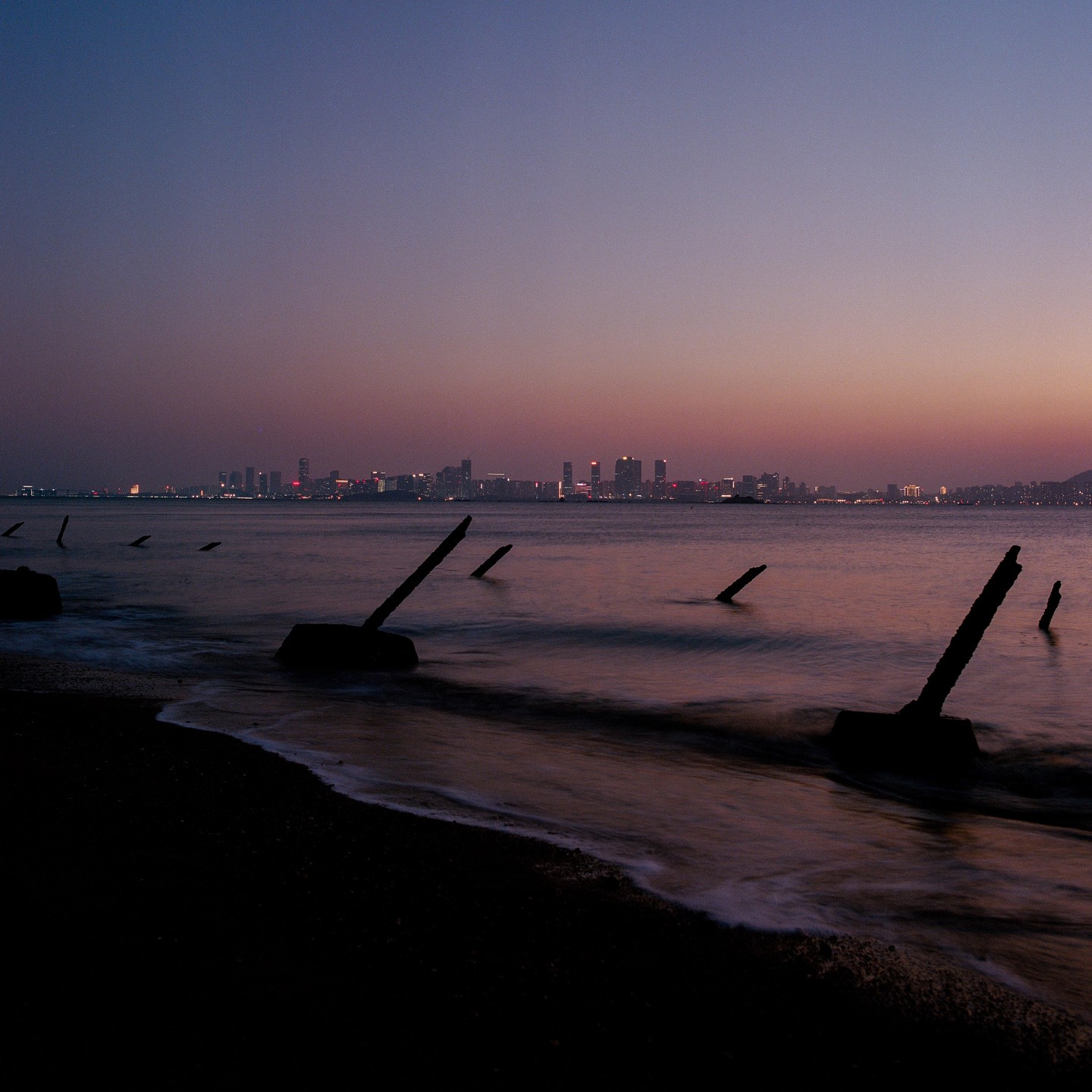 A city skyline is visible across the water at dusk, with several dark, angular structures emerging from the water in the foreground.