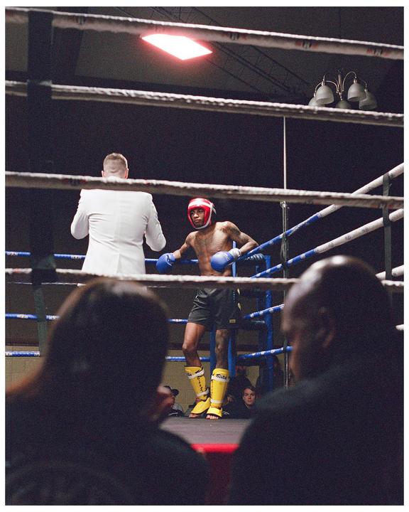 A boxer in a red headguard and blue gloves stands in a boxing ring with a man in a white suit.