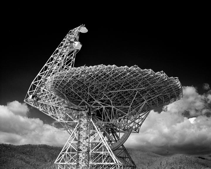 A large radio telescope stands against a dark sky with clouds and distant mountains.