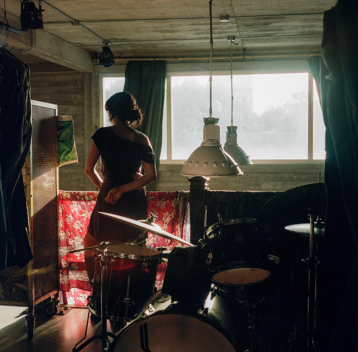 A woman stands with her back to the viewer in a room with drums and large windows.
