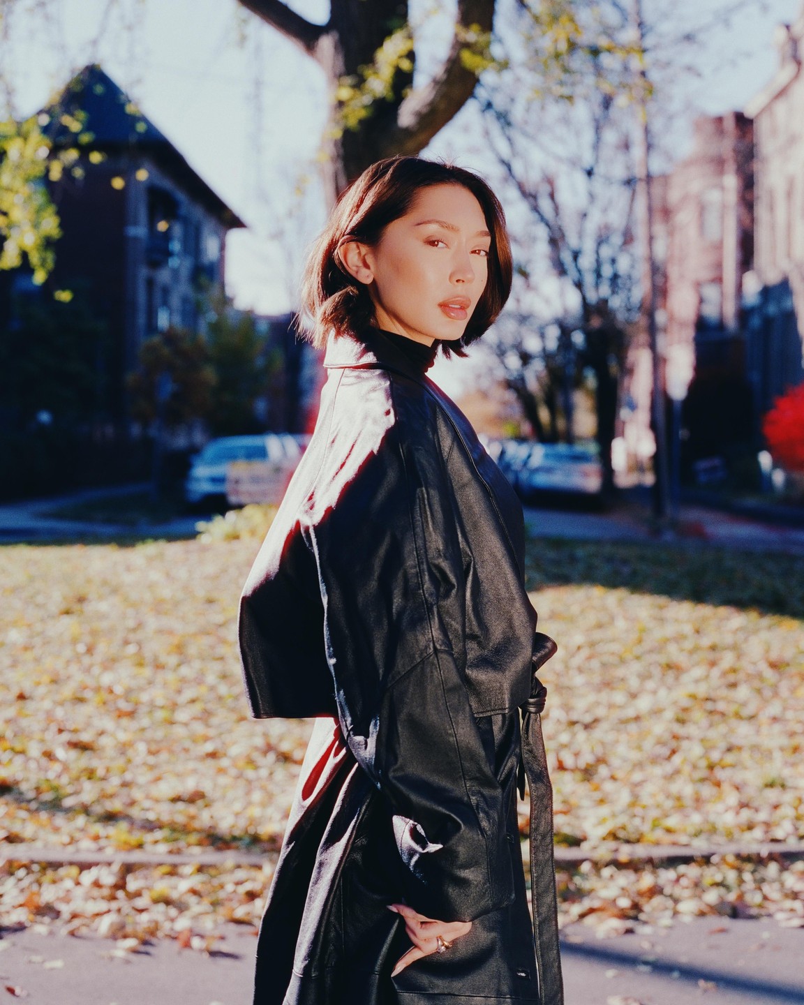 A woman in a black coat stands on a street with autumn leaves on the ground.