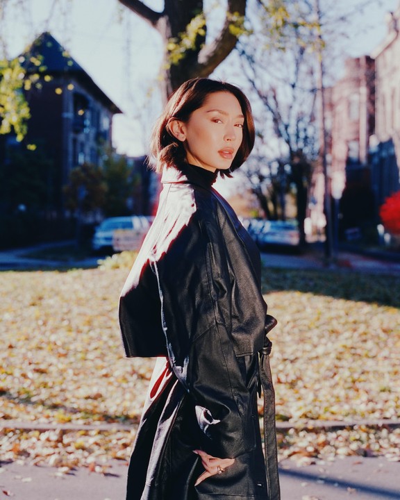 A woman in a black coat stands on a street with autumn leaves on the ground.