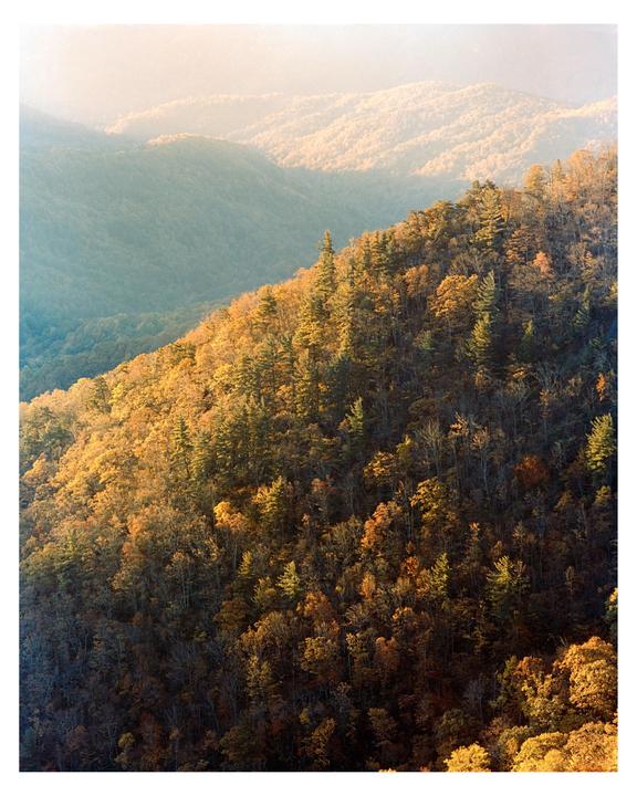 Sunlight illuminates a mountainside covered in autumn trees, with hazy mountains in the background.