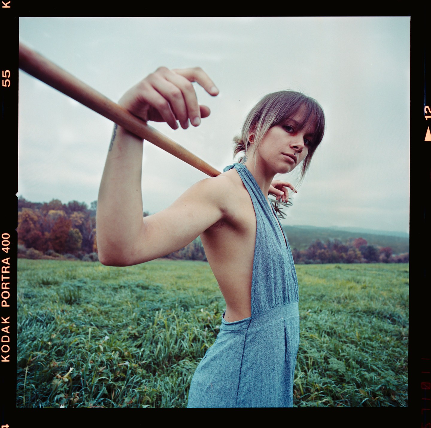 A woman in a blue dress holds a wooden stick over her shoulder in a grassy field with trees in the background.