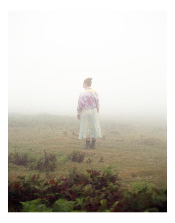 A woman stands in a field of low-lying plants, looking out into a thick fog.