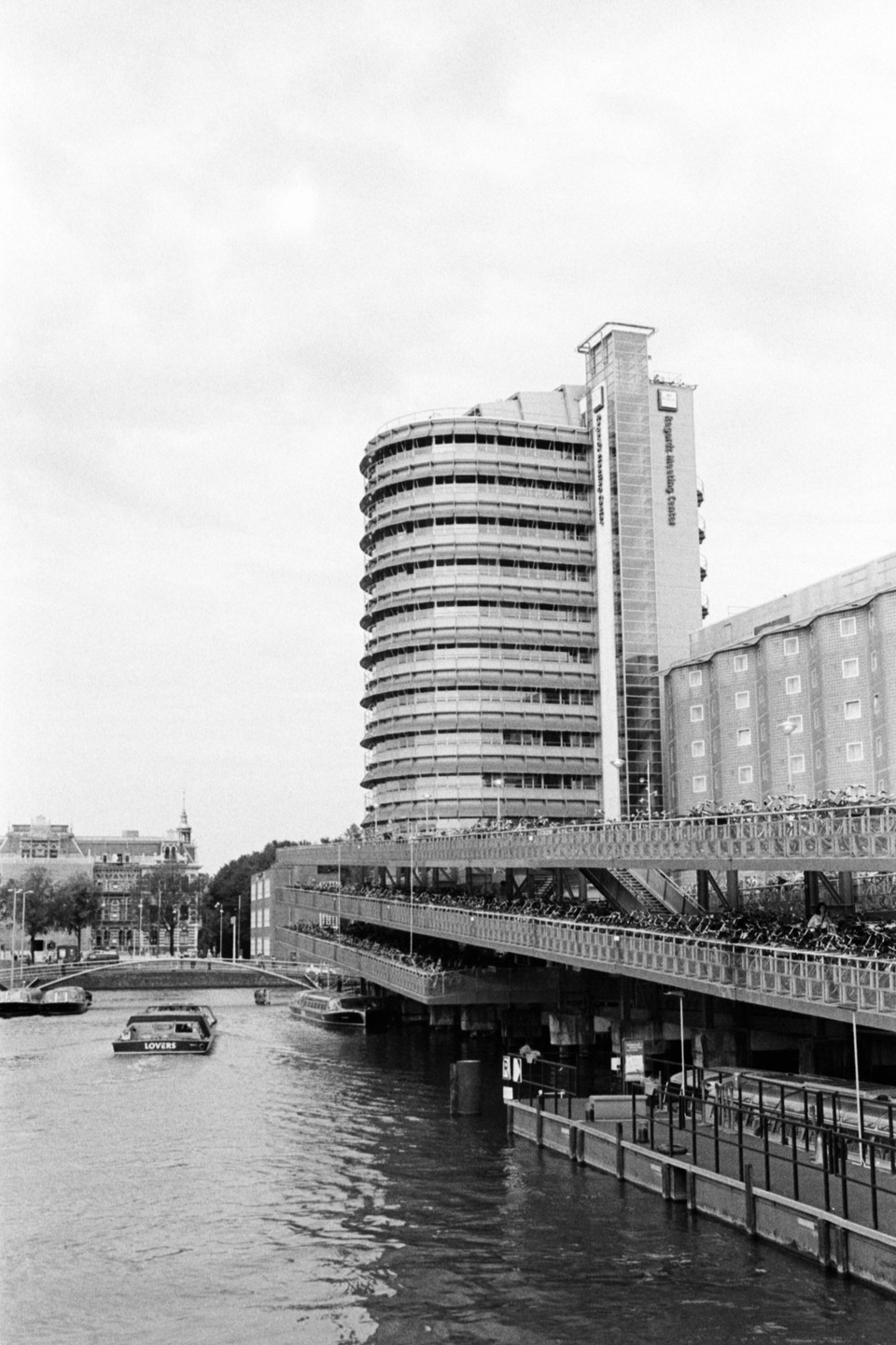 A canal with boats is in the foreground, with a large modern building and a multi-level bicycle parking structure in the background.