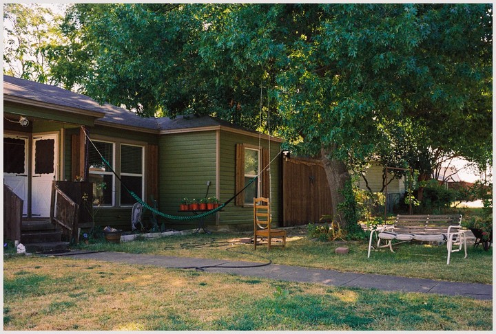 A green house with a porch and a large tree in the yard is shown on a sunny day.