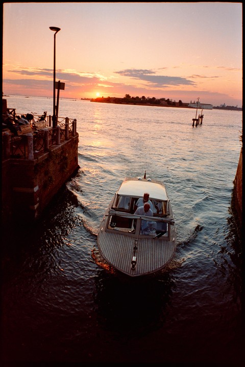 A boat with two men on board navigates a canal at sunset, with a distant island and buildings visible under a colorful sky.