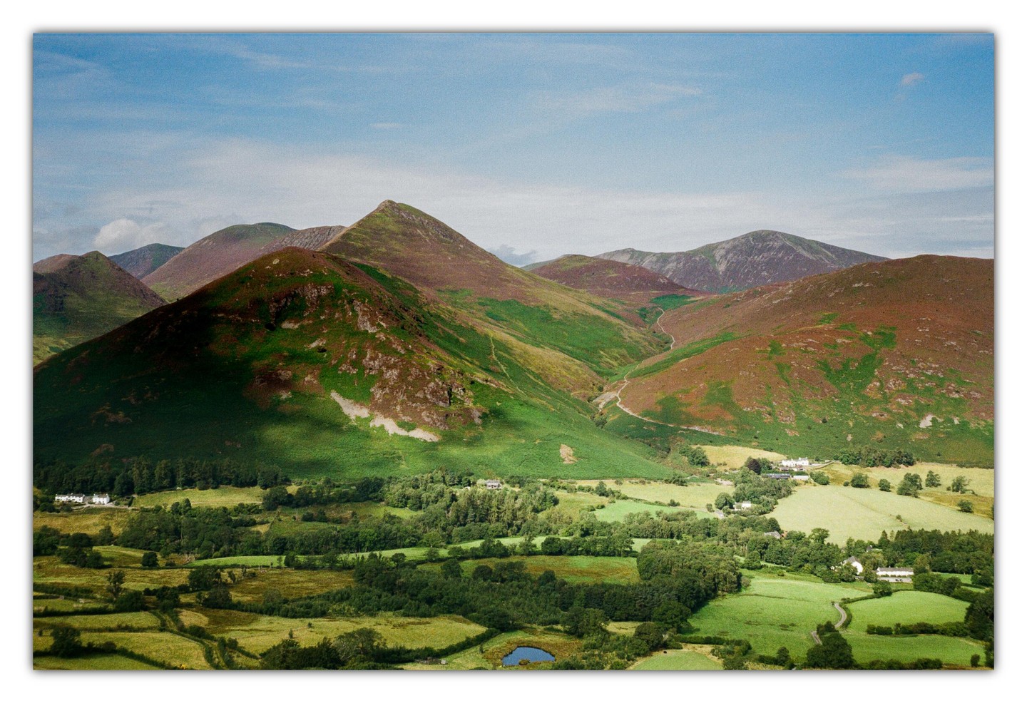 A panoramic landscape shows green and brown mountains overlooking a valley with fields, trees, and a small pond under a blue sky.