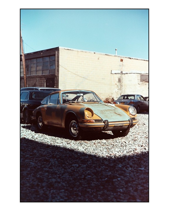 A rusty vintage car is parked on a gravel lot next to a building under a clear sky.