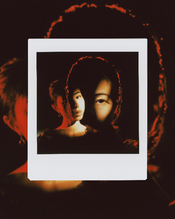 A woman's face is visible in a square instant print, with another woman's face visible in the background.