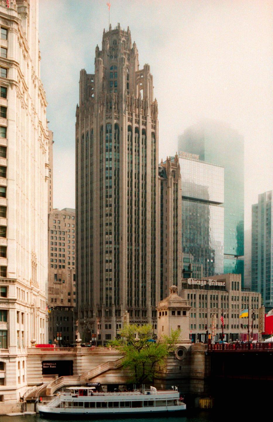 A tall, ornate building with a flag on top stands prominently among other city buildings, with a boat visible on the water below.