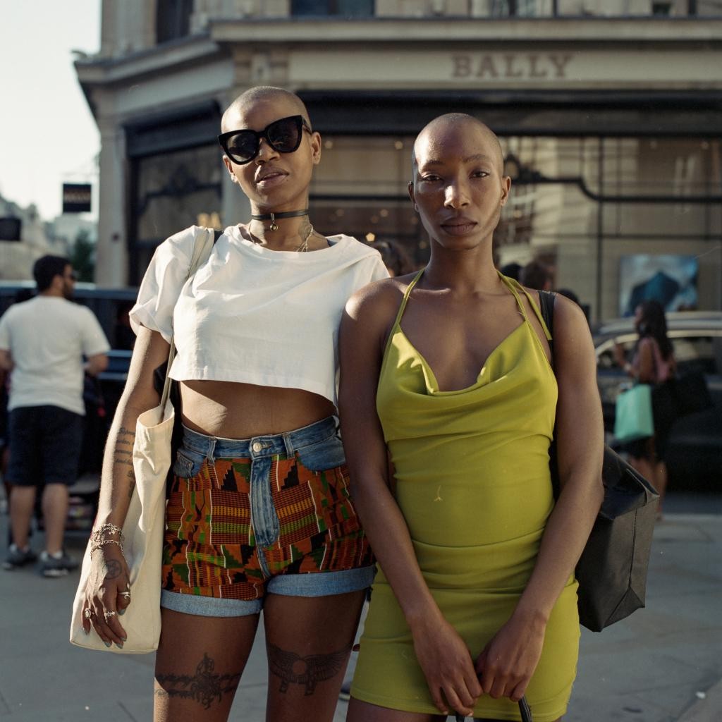 Two women with shaved heads stand on a street in front of a building with a "BALLY" sign.
