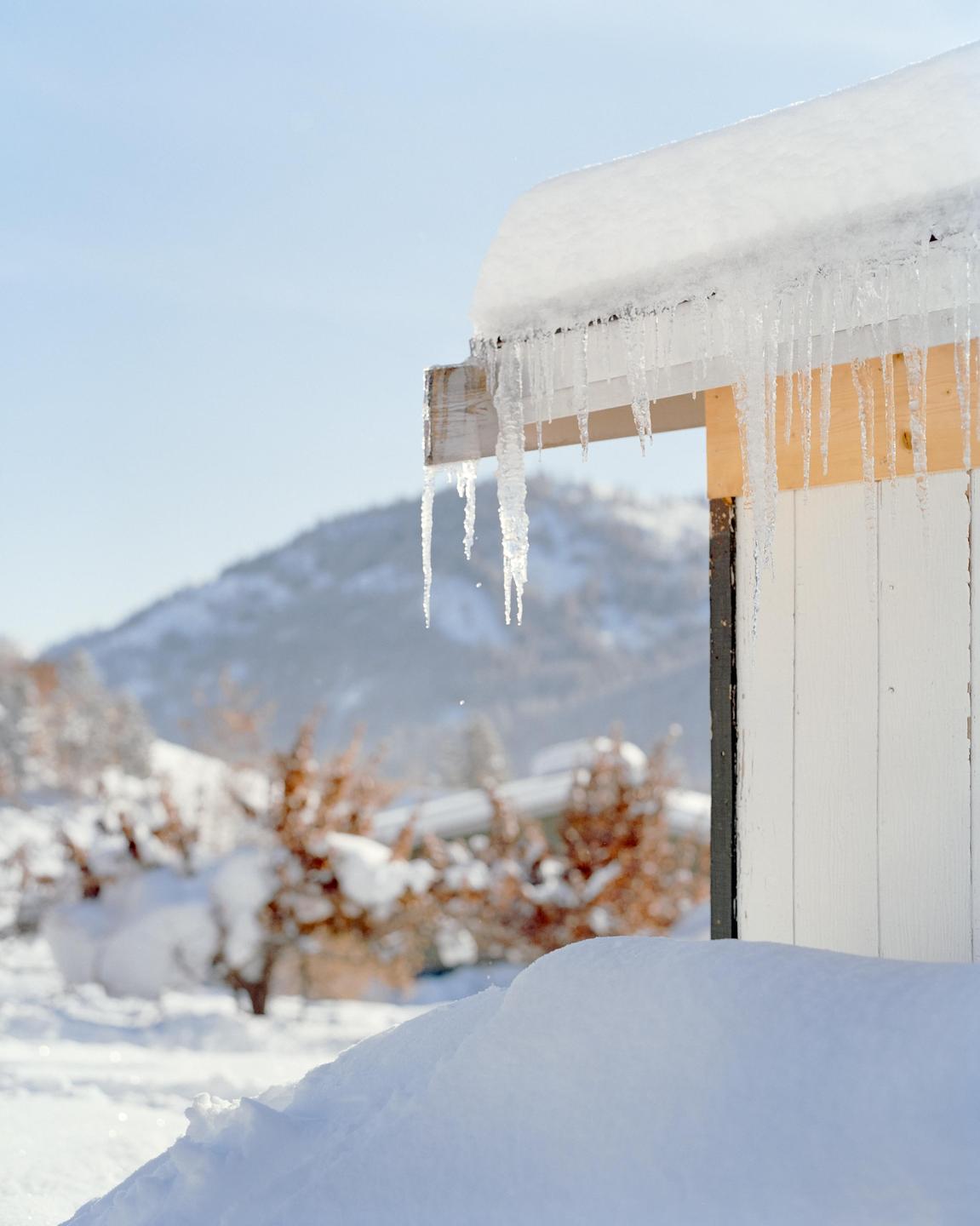 Icicles hang from the snow-covered roof of a white building with a snowy mountain in the background.