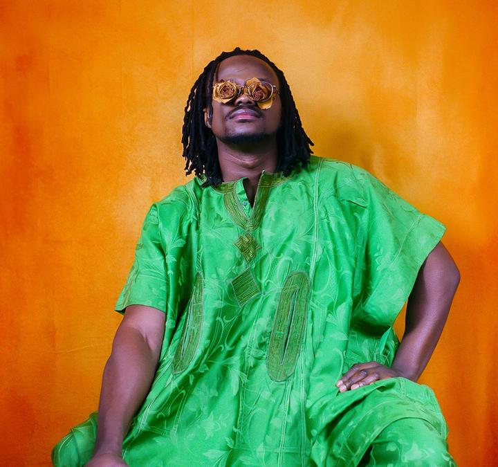 A man wearing a green tunic and glasses made of dried flowers sits against an orange background.