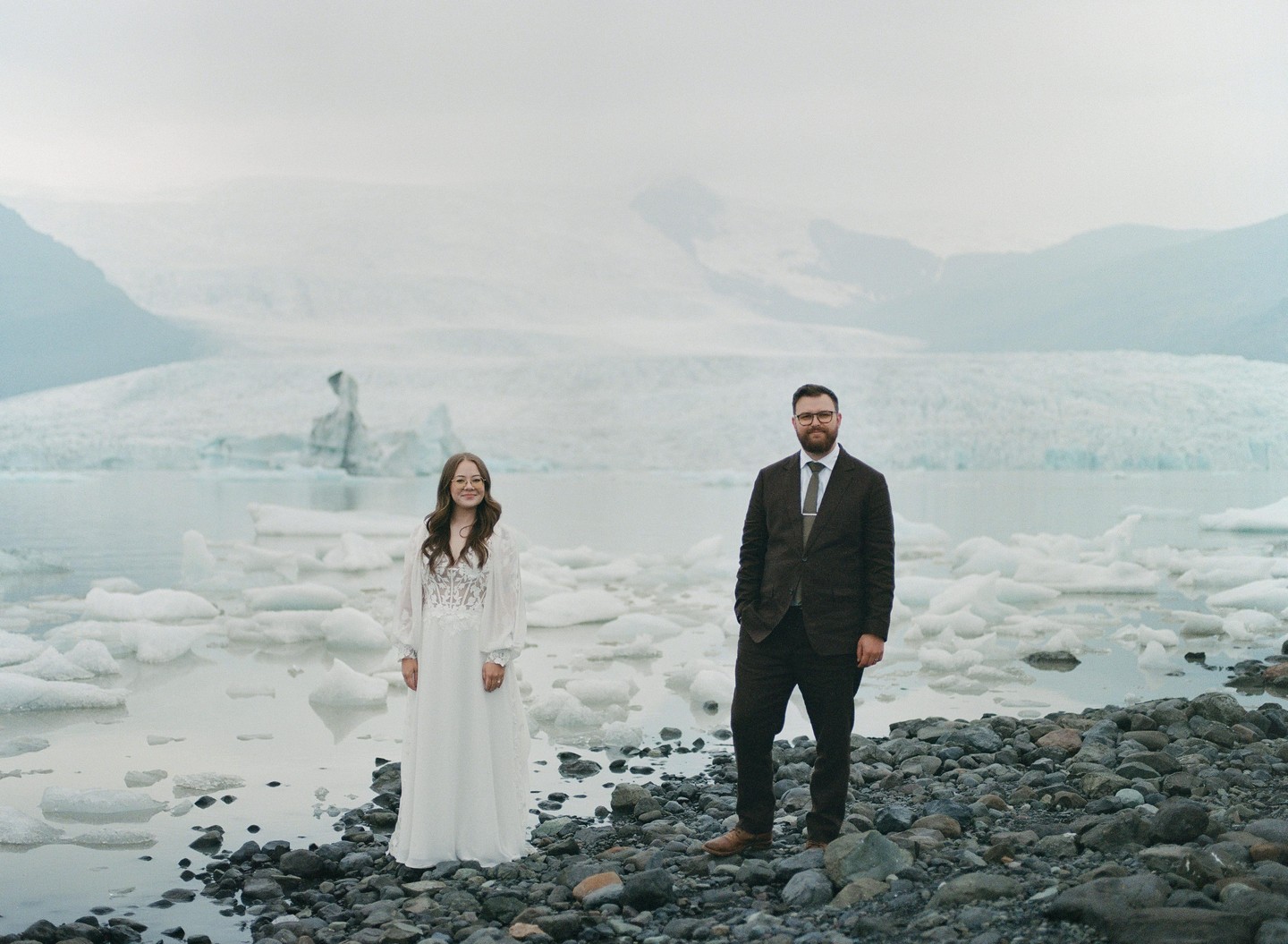 A couple stands on a rocky shore with icebergs floating in the water and a glacier in the background.