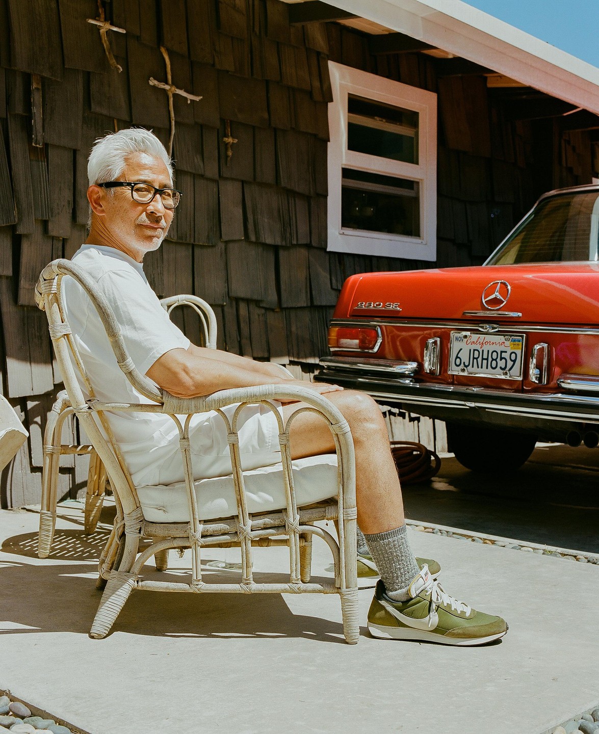 A man sits in a wicker chair next to a red car parked in front of a building with shingle siding.