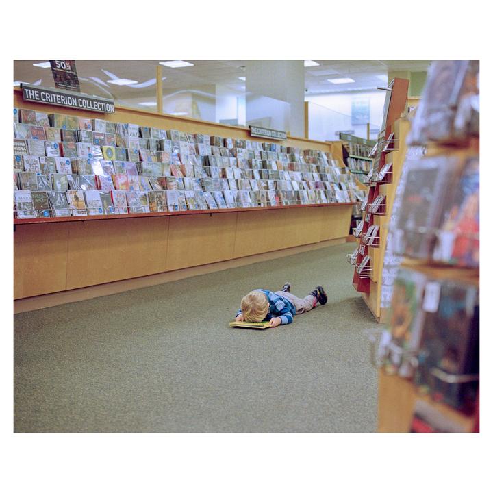 A child lies face down on a book on the carpeted floor of a bookstore.
