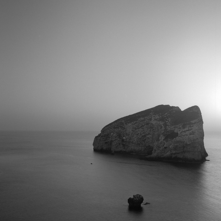 A large island rock formation sits in the ocean with a small boat nearby and a smaller rock in the foreground.