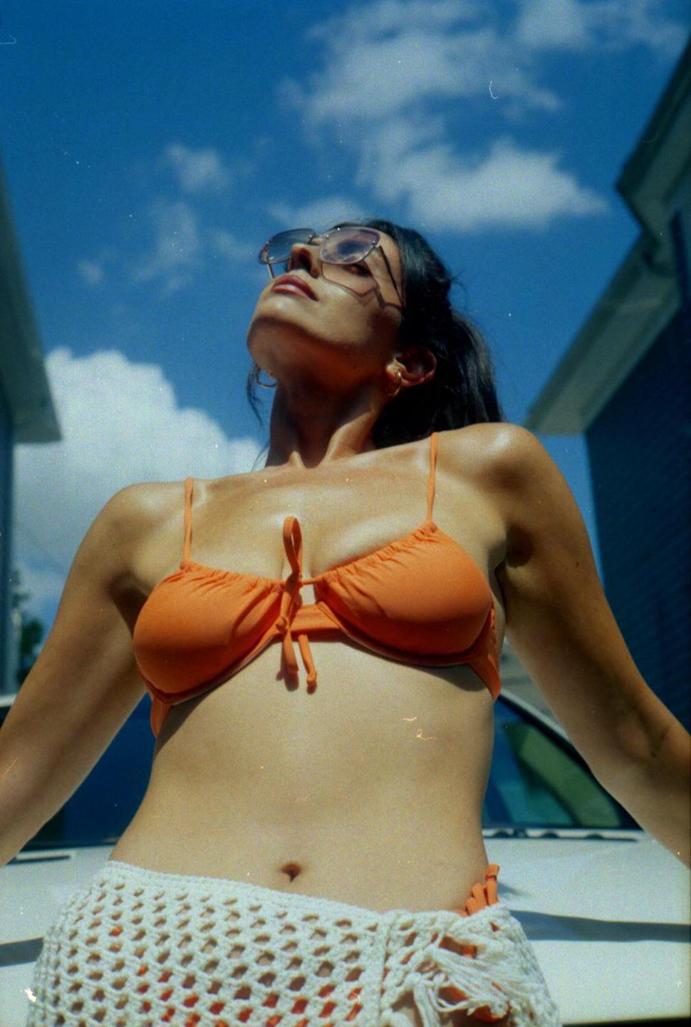 A woman wearing sunglasses, an orange bikini top, and a white crochet skirt looks up at the sky.