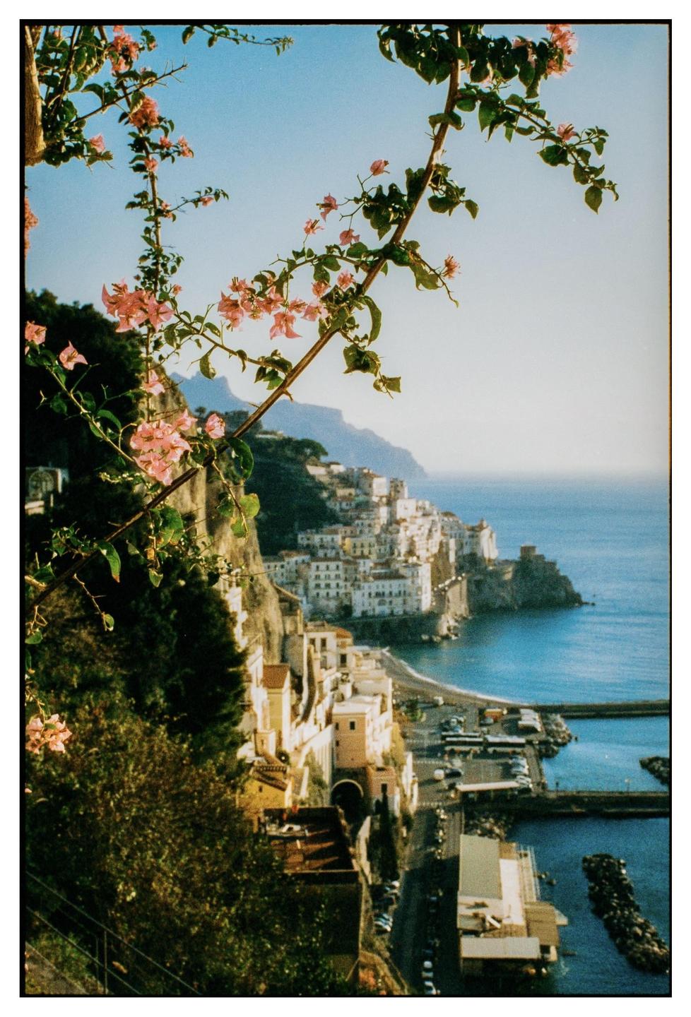 Pink bougainvillea flowers frame a view of a coastal town built into a cliffside overlooking the sea.