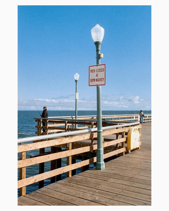 A man fishes from a wooden pier under a clear blue sky with a "pier closed at 10pm nightly" sign visible.