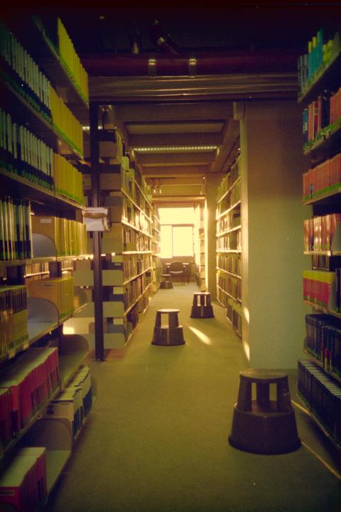 A long aisle in a library is lined with bookshelves and has three step stools on the carpeted floor, leading to a bright window at the end.