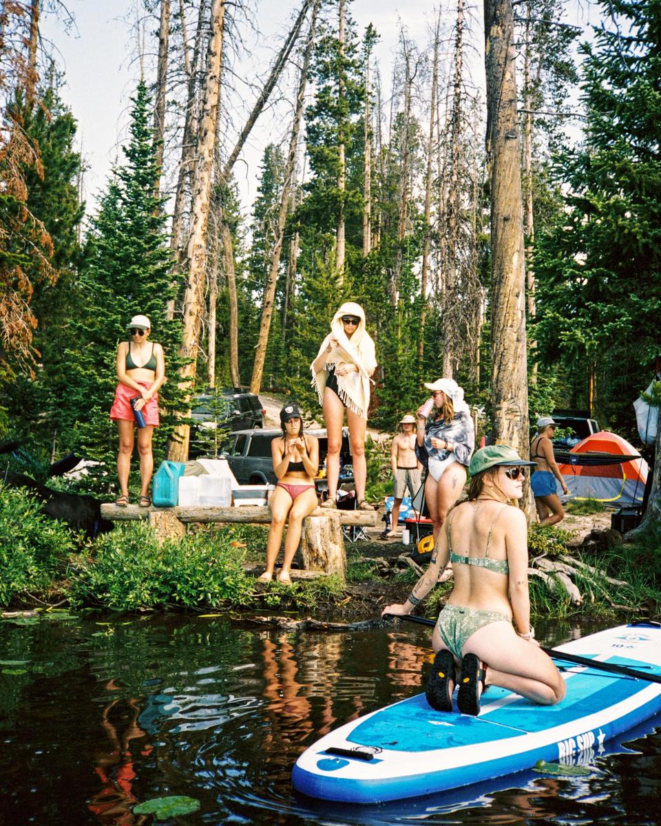 A group of women and a man are gathered by a body of water in a forest, with one woman kneeling on a paddleboard.